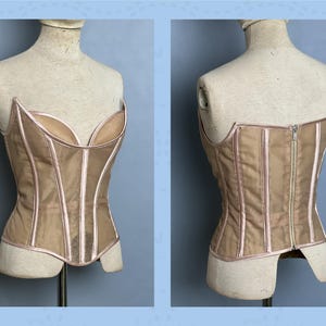 May include: A beige corset with a sweetheart neckline and a zipper closure. The corset is displayed on a mannequin and features vertical boning and pink trim. The front and back views are shown, highlighting the garment's construction and design.