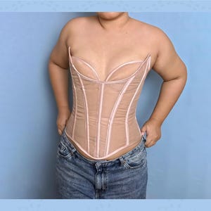May include: A beige corset with a sheer overlay and pink trim. The corset has a structured design with vertical boning and a sweetheart neckline. The garment is worn with blue denim jeans. The background is a light blue.