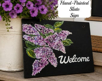 May include: A rectangular black slate sign featuring a hand-painted cluster of purple lilac flowers with green leaves and the white text "Welcome" in a script font. The sign has two circular holes at the top for hanging purposes.