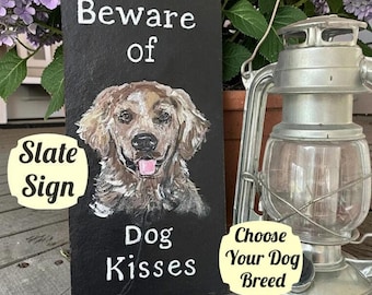 May include: A rectangular black slate sign featuring a hand-painted portrait of a golden-colored dog with its tongue out. The sign displays the white text "Beware of" at the top and "Dog Kisses" at the bottom, with additional text overlays.