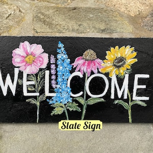 May include: A black slate sign with the word "WELCOME" painted in white. The sign is decorated with painted flowers in pink, blue, yellow, and green.