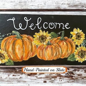Puede incluir: Un letrero rectangular de pizarra negra con el texto Welcome pintado a mano en blanco en la parte superior. El dise&ntilde;o incluye dos grandes calabazas naranjas, tres girasoles amarillos y hojas verdes pintadas en el centro de la superficie.