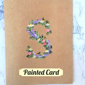 May include: A brown card with a painted floral letter S. The letter is made of purple lavender, pink roses, white daisies, and green leaves. There are small yellow bees flying around the letter. The card has the text "Painted Card" printed on it.