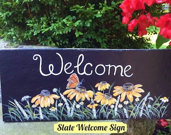 May include: Black slate welcome sign with a hand-painted design of yellow flowers, green grass, and a butterfly. The sign reads "Welcome".