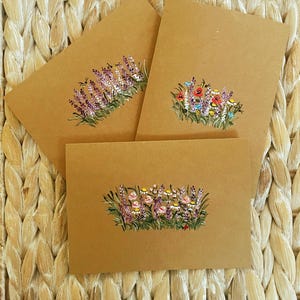 May include: Three rectangular brown kraft paper cards, each featuring a hand-painted botanical illustration of wildflowers and greenery. The designs include clusters of purple, red, pink, and white flowers with green stems and small bees.