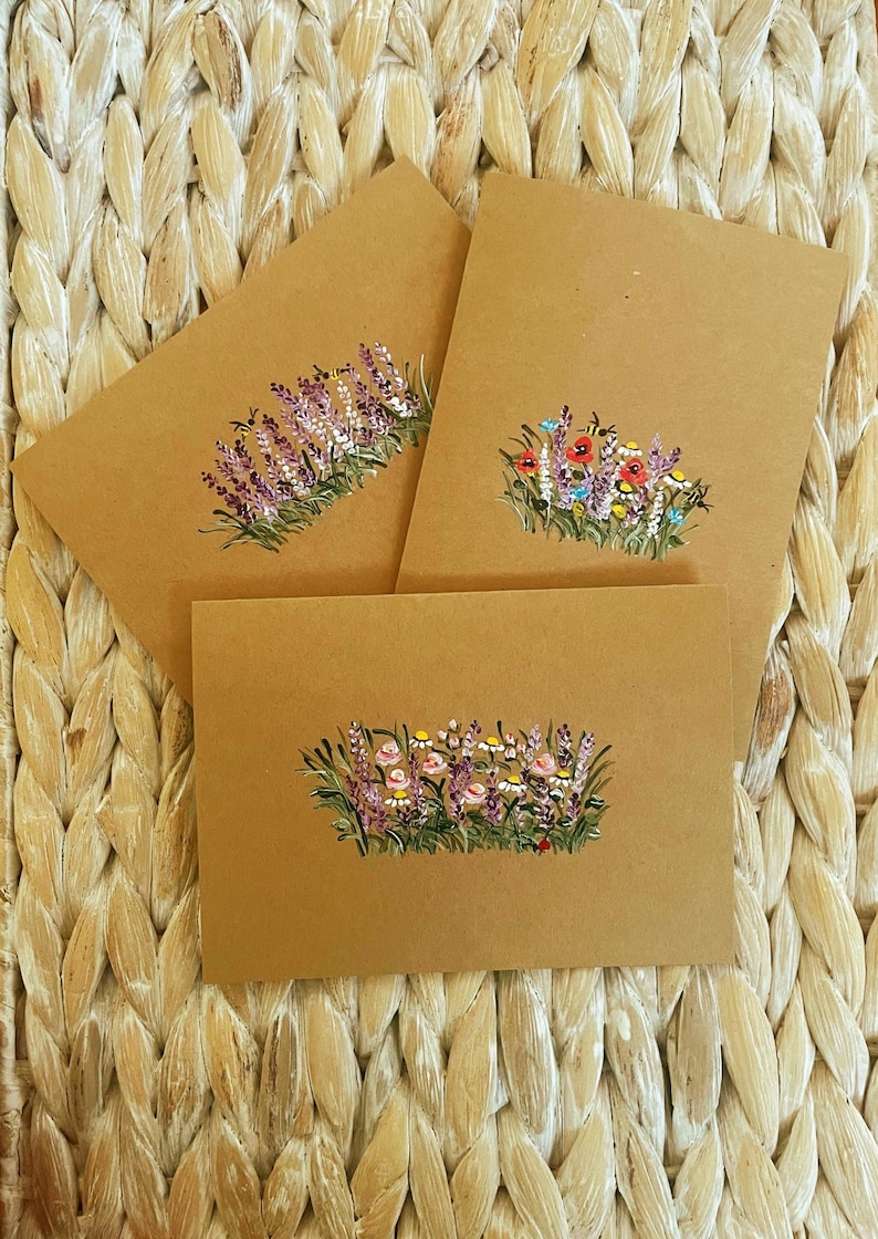 May include: Three rectangular brown kraft paper cards, each featuring a hand-painted botanical illustration of wildflowers and greenery. The designs include clusters of purple, red, pink, and white flowers with green stems and small bees.