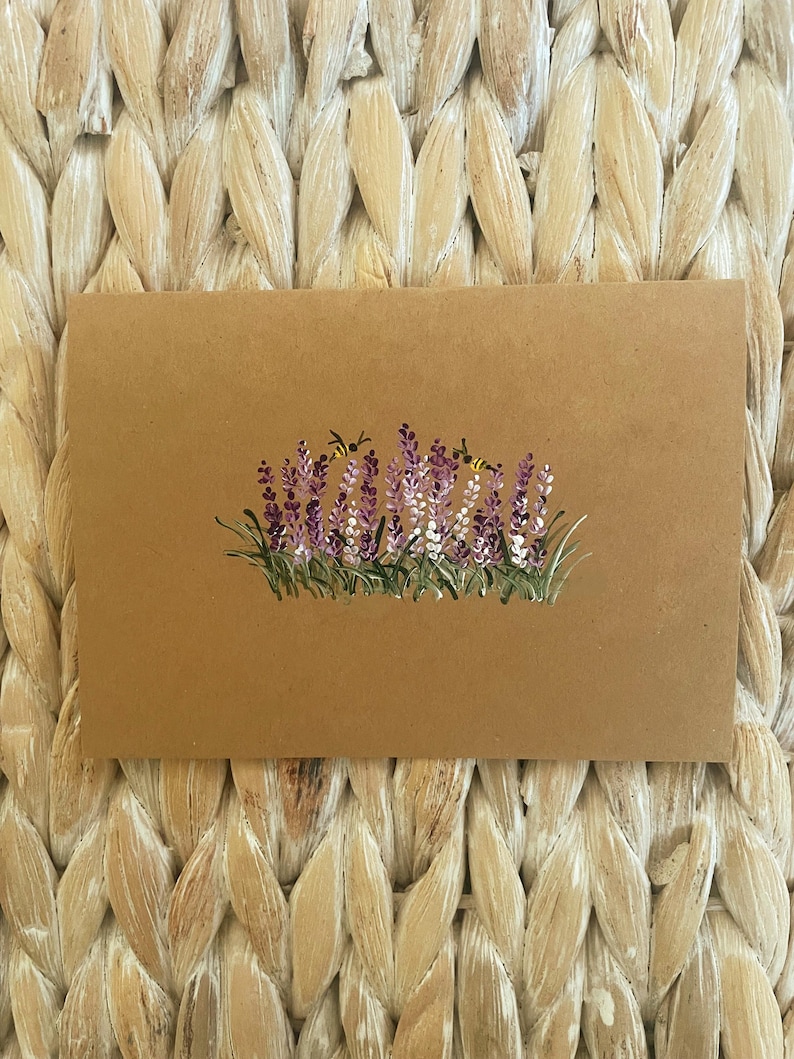 May include: A rectangular tan card featuring a hand-painted illustration of a cluster of purple and white lavender flowers with green stems and leaves. Two small yellow and black bees are depicted hovering near the top of the floral arrangement.