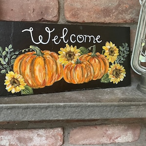 Puede incluir: Un letrero rectangular de pizarra negra con la palabra Welcome pintada a mano en letra cursiva blanca. El dise&ntilde;o incluye tres calabazas naranjas y cuatro girasoles amarillos con hojas verdes dispuestos en la parte inferior.