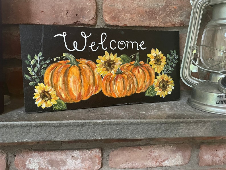 Puede incluir: Un letrero rectangular de pizarra negra con la palabra Welcome pintada a mano en letra cursiva blanca. El dise&ntilde;o incluye tres calabazas naranjas y cuatro girasoles amarillos con hojas verdes dispuestos en la parte inferior.