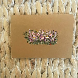 May include: A rectangular tan card featuring a hand-painted illustration of a wildflower meadow. The artwork displays purple lavender stalks, small white daisies with yellow centers, and soft pink blossoms nestled among green grass blades.