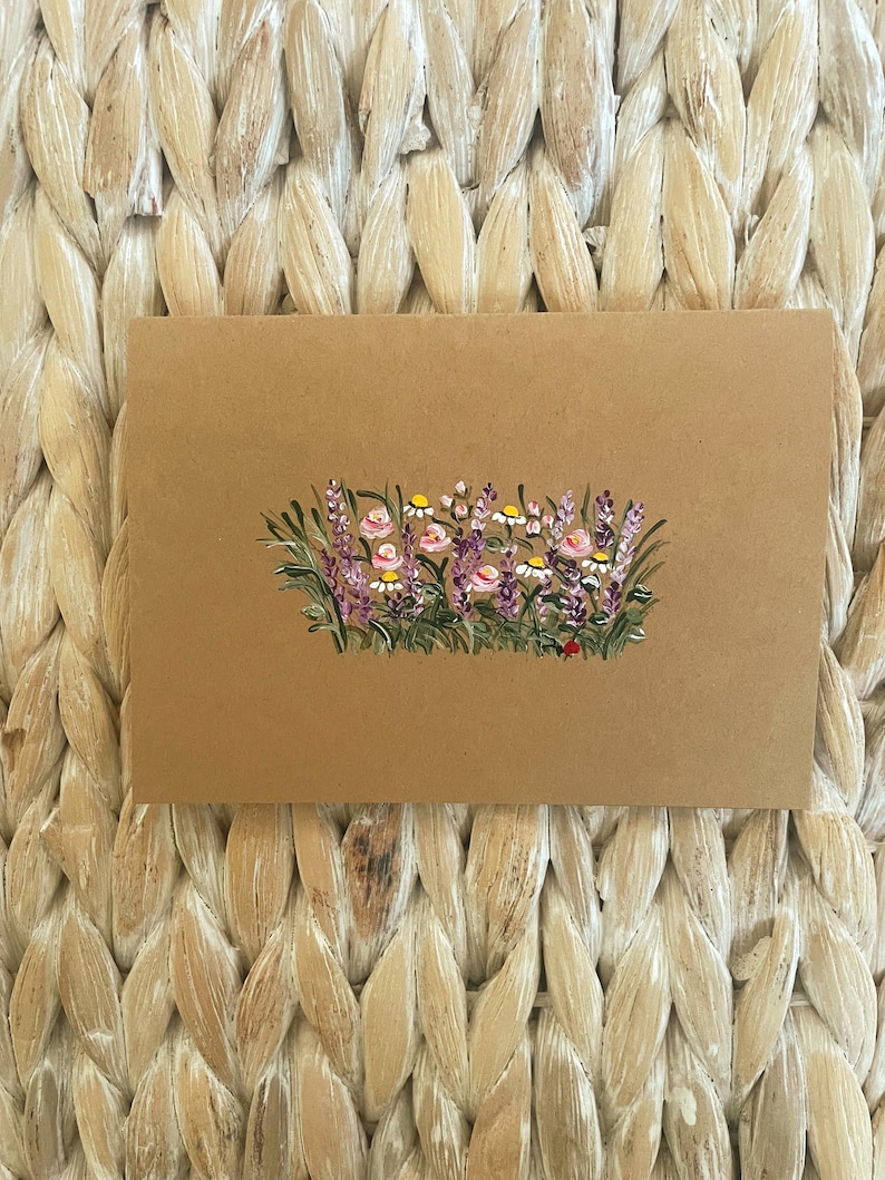 May include: A rectangular tan card featuring a hand-painted illustration of a wildflower meadow. The artwork displays purple lavender stalks, small white daisies with yellow centers, and soft pink blossoms nestled among green grass blades.