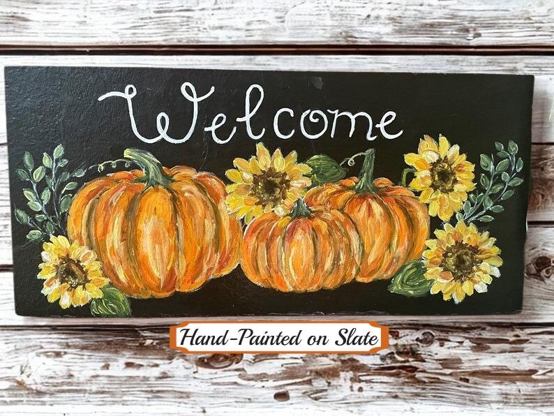 Puede incluir: Un letrero rectangular de pizarra negra con el texto Welcome pintado a mano en blanco en la parte superior. El dise&ntilde;o incluye dos grandes calabazas naranjas, tres girasoles amarillos y hojas verdes pintadas en el centro de la superficie.