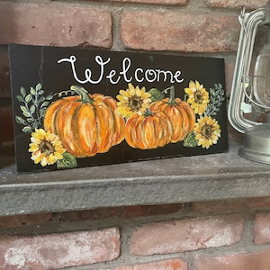 Puede incluir: Un letrero rectangular negro con el texto Welcome pintado a mano en blanco, decorado con calabazas naranjas, girasoles amarillos y hojas verdes. El objeto se encuentra sobre una repisa junto a una linterna de metal plateado.