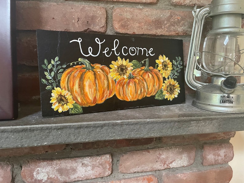 Puede incluir: Un letrero rectangular negro con el texto Welcome pintado a mano en blanco, decorado con calabazas naranjas, girasoles amarillos y hojas verdes. El objeto se encuentra sobre una repisa junto a una linterna de metal plateado.