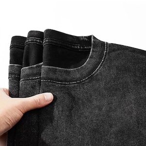 May include: Close-up of several black t-shirts with a faded, vintage wash. The shirts are stacked, showing the crew neck and shoulder seams. The fabric has a textured appearance, and the color is a deep, charcoal black.