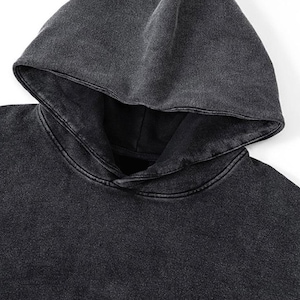 May include: Close-up of a dark gray hoodie with a hood. The fabric has a textured, washed appearance. The hood is folded down, revealing the neckline. The hoodie is made of a soft material.