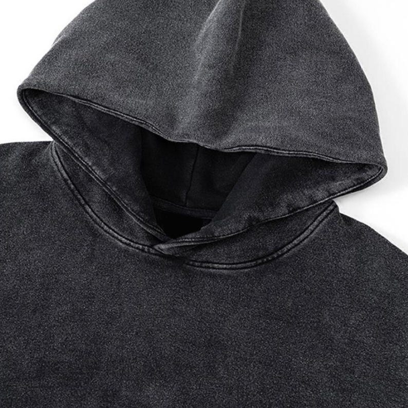 May include: Close-up of a dark gray hoodie with a hood. The fabric has a textured, washed appearance. The hood is folded down, revealing the neckline. The hoodie is made of a soft material.