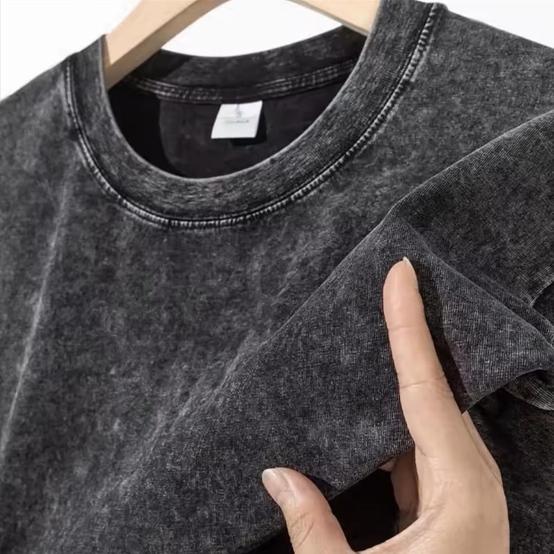 May include: A dark gray, acid-wash crew neck sweatshirt. The fabric has a mottled, textured appearance. The garment is hanging on a wooden hanger. The collar and seams are visible.