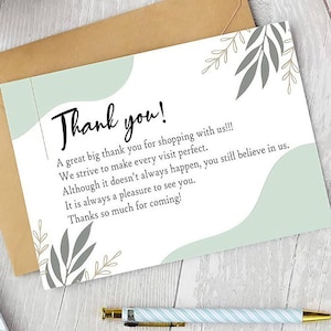 May include: A thank you card with the words "Thank you!" in black script. The card has a light green and gray leaf design. The card is on top of a brown envelope.