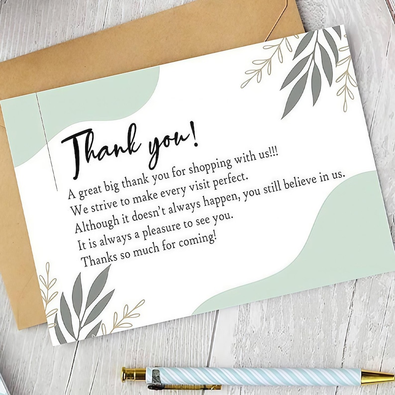 May include: A thank you card with the words "Thank you!" in black script. The card has a light green and gray leaf design. The card is on top of a brown envelope.