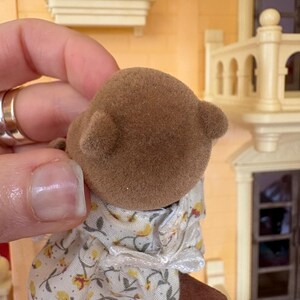 May include: A brown flocked bear figurine wearing a floral print dress with yellow flowers and green leaves. The bear has small, rounded ears. The background includes a miniature house with a window box of colorful flowers.