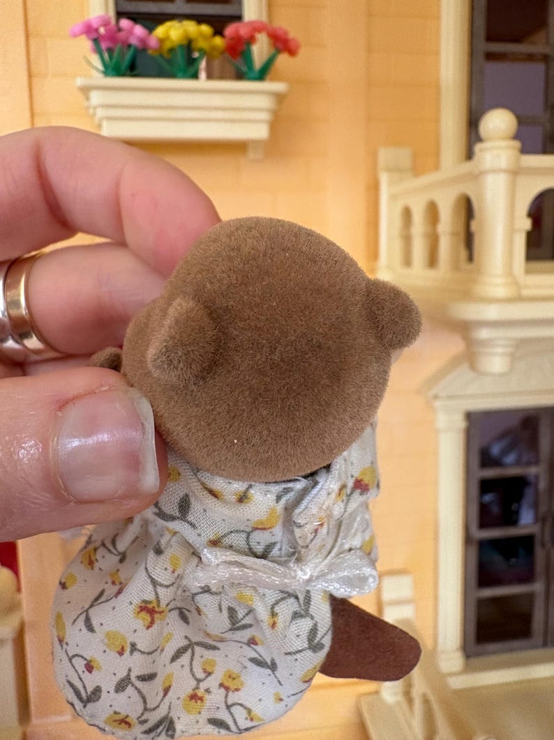 May include: A brown flocked bear figurine wearing a floral print dress with yellow flowers and green leaves. The bear has small, rounded ears. The background includes a miniature house with a window box of colorful flowers.