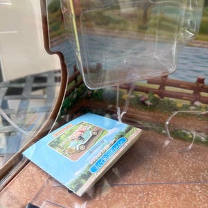 May include: A blue and white booklet with Japanese text and an illustration of a vintage car carrying a person. The booklet is inside a clear plastic container. The background shows a scenic landscape with a lake and a wooden fence.