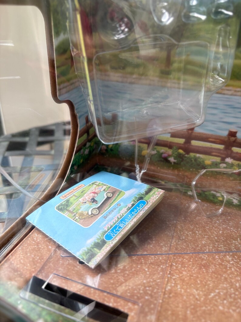 May include: A blue and white booklet with Japanese text and an illustration of a vintage car carrying a person. The booklet is inside a clear plastic container. The background shows a scenic landscape with a lake and a wooden fence.