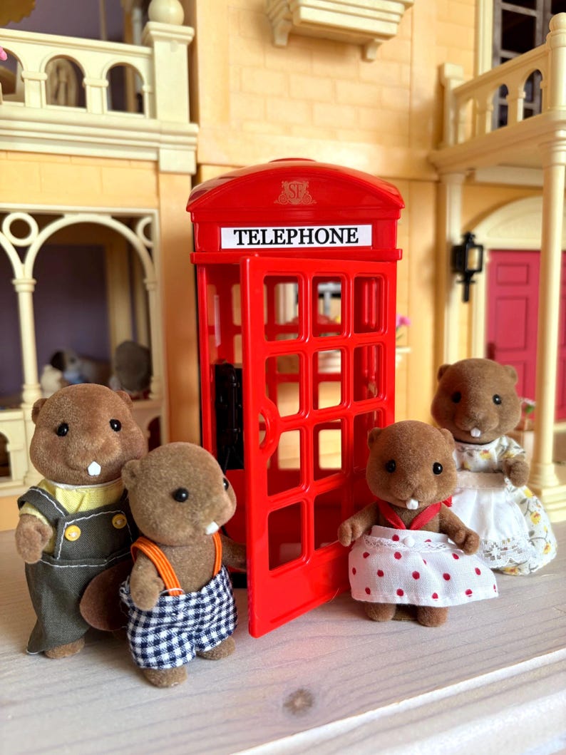 May include: A miniature scene featuring a red telephone booth with the word "TELEPHONE" on top. Four brown beaver figurines are dressed in various outfits, standing near the booth. The scene is set in front of a dollhouse.