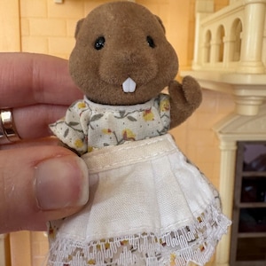 May include: A brown beaver figurine wearing a floral print dress with a white apron. The apron has a lace trim. The figurine has black eyes and a white tooth. The background includes a dollhouse.