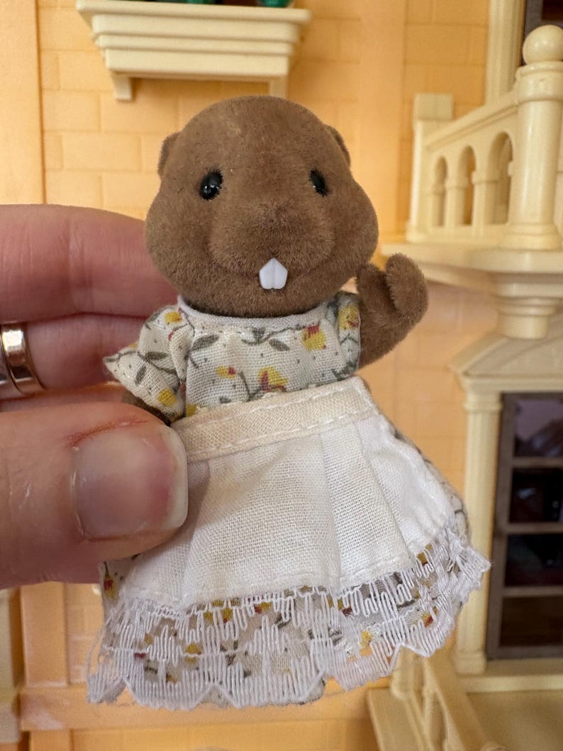 May include: A brown beaver figurine wearing a floral print dress with a white apron. The apron has a lace trim. The figurine has black eyes and a white tooth. The background includes a dollhouse.