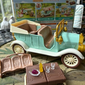 May include: A vintage-style toy car in mint green and cream, with brown interior and white wheels. Accessories include a brown picnic table with a bottle, grapes, and glasses, and a brown bench. The box reads "Sylvanian Families".