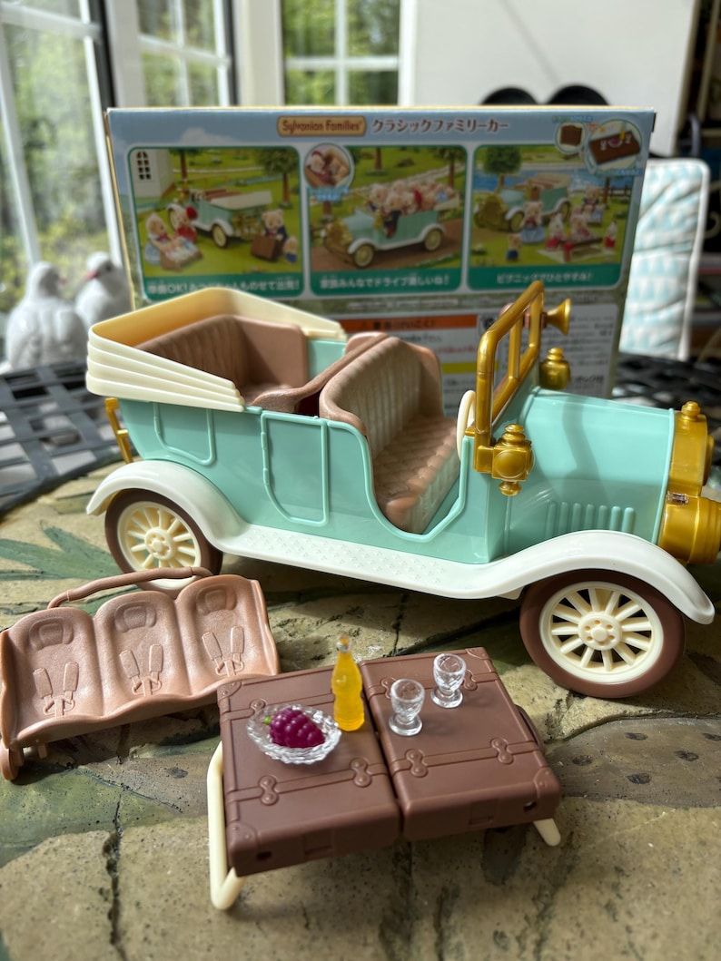 May include: A vintage-style toy car in mint green and cream, with brown interior and white wheels. Accessories include a brown picnic table with a bottle, grapes, and glasses, and a brown bench. The box reads "Sylvanian Families".