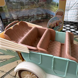 May include: A vintage-style toy car in pastel blue and white, with brown seating. The car has a detailed design, including a white fender and a cream-colored wheel. The packaging behind the car features a scenic illustration and Japanese text.