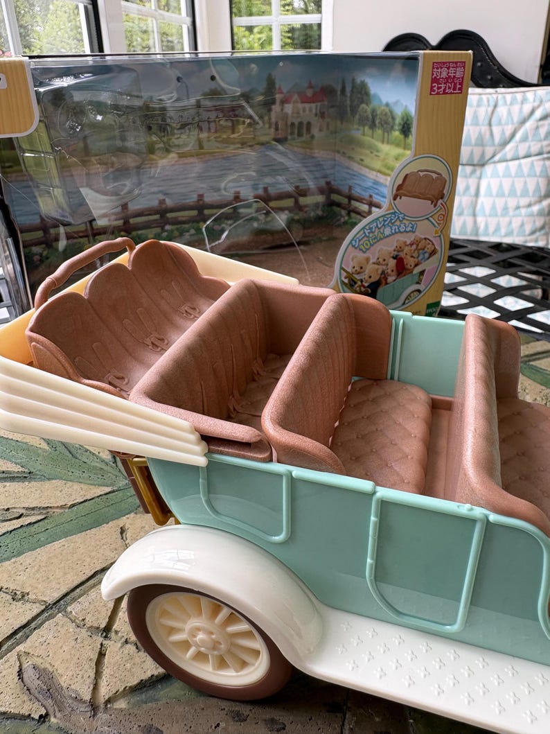 May include: A vintage-style toy car in pastel blue and white, with brown seating. The car has a detailed design, including a white fender and a cream-colored wheel. The packaging behind the car features a scenic illustration and Japanese text.
