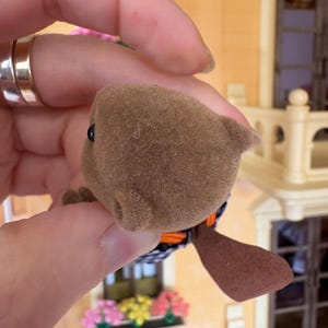 May include: A brown, plush toy beaver with a black and white patterned body, orange accents, and a flat, brown tail. The toy has a small, black eye and is held in a hand.