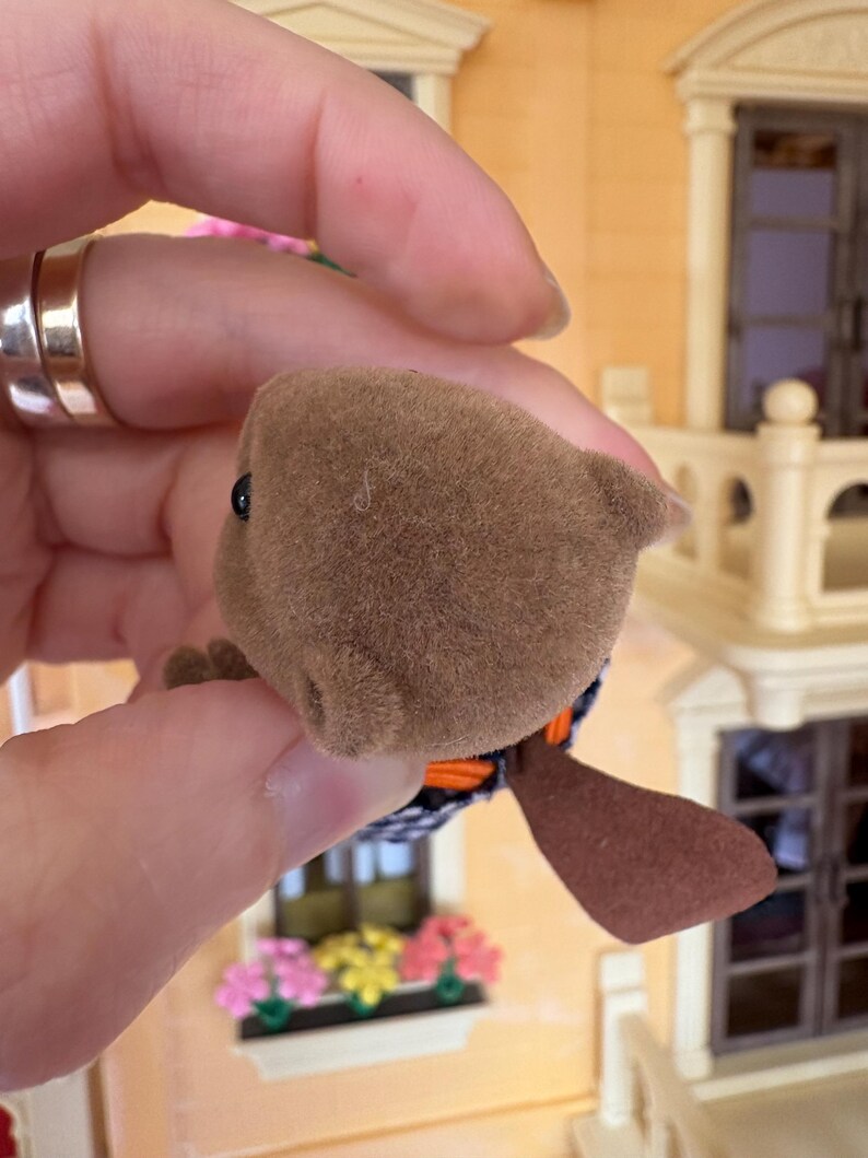 May include: A brown, plush toy beaver with a black and white patterned body, orange accents, and a flat, brown tail. The toy has a small, black eye and is held in a hand.