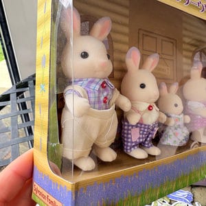May include: A Sylvanian Families Lavender Rabbit family set in a clear plastic package. The set includes five white rabbit figures wearing colorful outfits. The packaging features lavender and Japanese text.