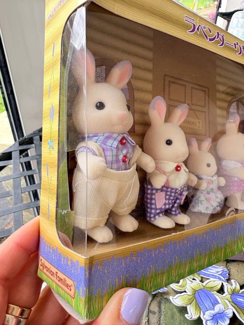 May include: A Sylvanian Families Lavender Rabbit family set in a clear plastic package. The set includes five white rabbit figures wearing colorful outfits. The packaging features lavender and Japanese text.