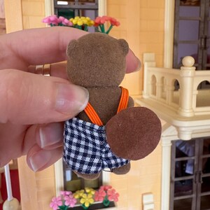 May include: A brown toy beaver figure with a checkered blue and white shorts and orange suspenders. The beaver has a large, flat, brown tail. The figure is held in front of a miniature building with a window box of flowers.