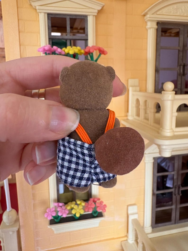May include: A brown toy beaver figure with a checkered blue and white shorts and orange suspenders. The beaver has a large, flat, brown tail. The figure is held in front of a miniature building with a window box of flowers.