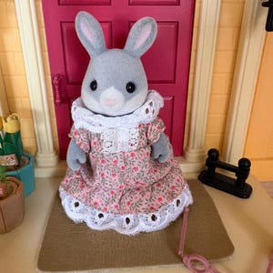 Może przedstawiać: Szara figurka kr&oacute;lika w r&oacute;żowej sukience w kwiaty z białym koronkowym wykończeniem i białym koronkowym kołnierzem. Kr&oacute;lik stoi przed r&oacute;żowymi drzwiami. R&oacute;żowe okulary są przed kr&oacute;likiem.