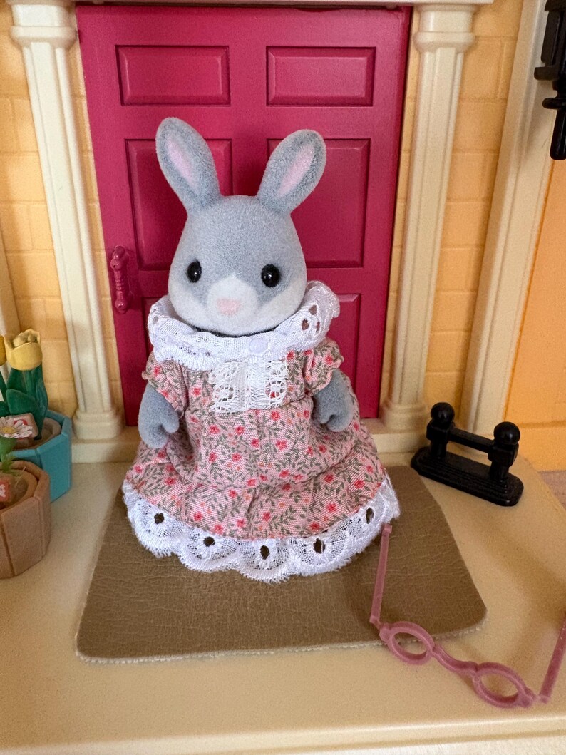 Może przedstawiać: Szara figurka kr&oacute;lika w r&oacute;żowej sukience w kwiaty z białym koronkowym wykończeniem i białym koronkowym kołnierzem. Kr&oacute;lik stoi przed r&oacute;żowymi drzwiami. R&oacute;żowe okulary są przed kr&oacute;likiem.