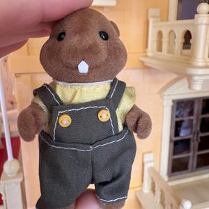 May include: A brown plush beaver figurine wearing a yellow shirt and dark green overalls with yellow buttons. The beaver has black eyes and a single white tooth. The figurine is part of a miniature toy set.