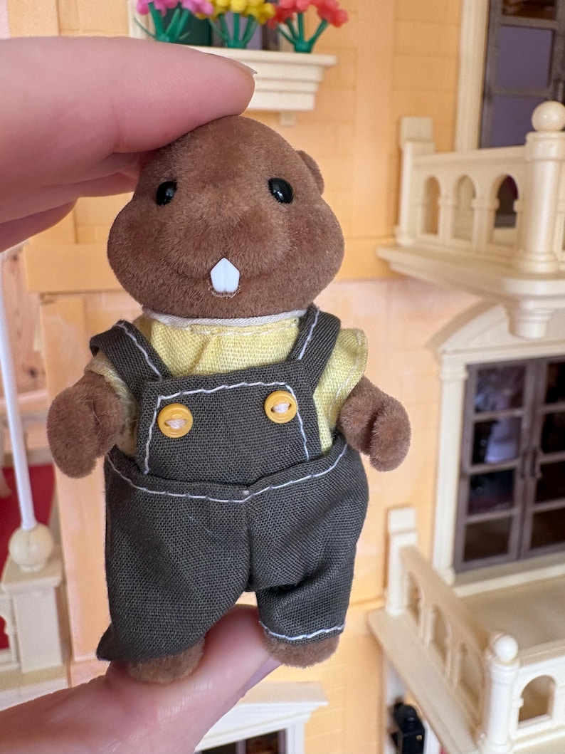 May include: A brown plush beaver figurine wearing a yellow shirt and dark green overalls with yellow buttons. The beaver has black eyes and a single white tooth. The figurine is part of a miniature toy set.