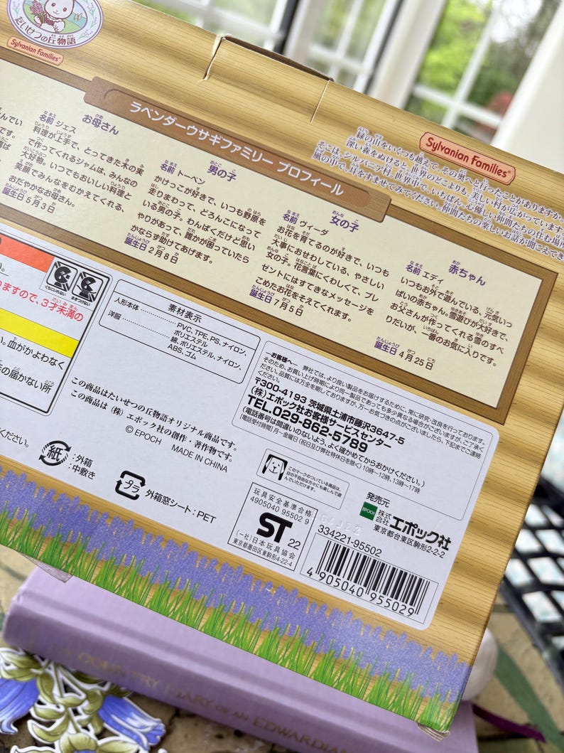 May include: A close-up of a box featuring Japanese text and the "Sylvanian Families" logo. The box has a lavender and wood-grain design. The text describes the Lavender Rabbit Family, including character names and birthdays.