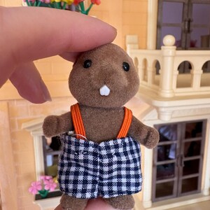 May include: A brown toy beaver figure with black eyes and a white tooth. It wears blue and white checkered shorts with orange suspenders. The figure is held in front of a building with a balcony and flowers.