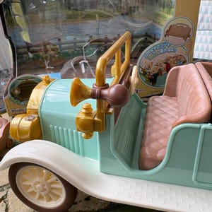 May include: A vintage-style toy car in pastel colors. The car features a light blue body, white fenders, and brown accents. It has a tan interior and a gold-colored horn and steering column. The toy is packaged with a scenic backdrop.