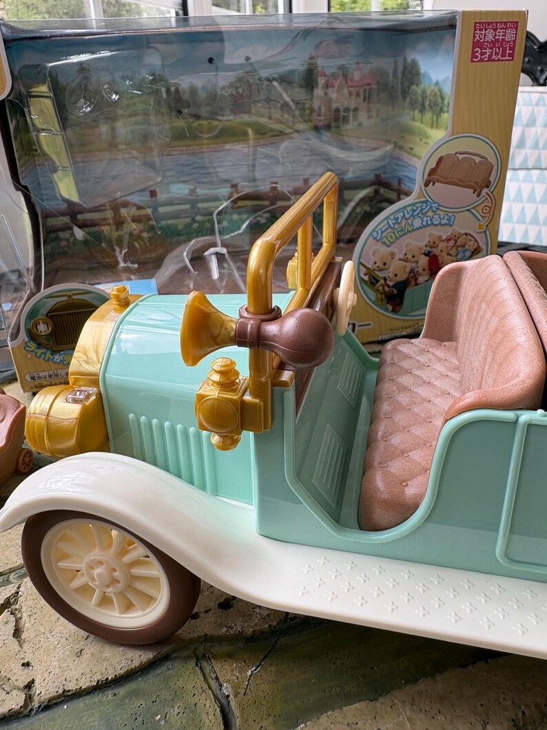 May include: A vintage-style toy car in pastel colors. The car features a light blue body, white fenders, and brown accents. It has a tan interior and a gold-colored horn and steering column. The toy is packaged with a scenic backdrop.