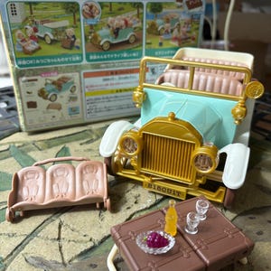 May include: A toy set featuring a vintage-style car in light blue and gold, with a matching bench and picnic table. Accessories include a bottle, glasses, and a plate of grapes. The box reads "Sylvanian Families".